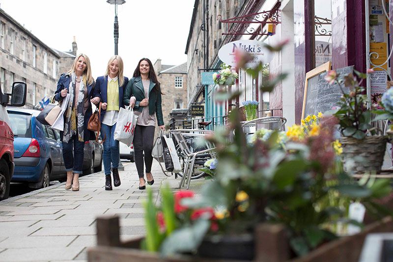Three women linking arms and smiling, holding many shopping bags and walking through Edinburgh