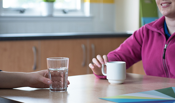 Close up of two people sat at a table, one has a glass of water and the other a mug