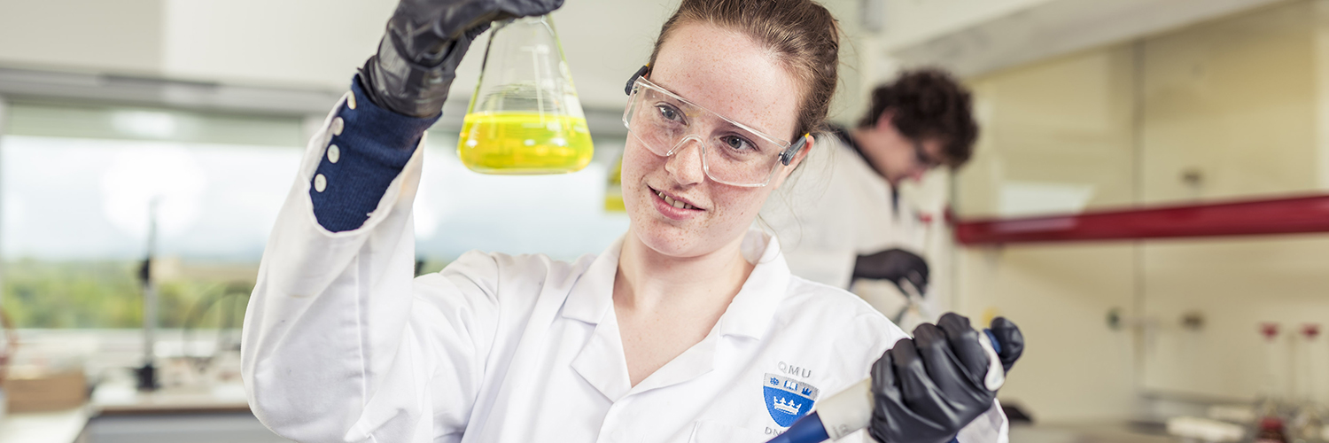 A QMU student wearing PPE and holding up a beaker of yellow liquid
