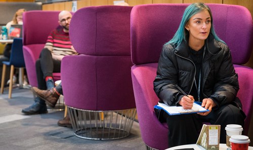 Student with vibrant blue hair sat on purple pod seat in QMU Atrium
